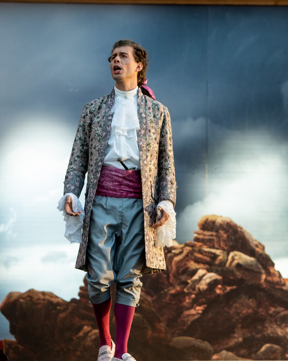 Maximilian Vogler singt auf der Opernbühne im historischen Kostüm vor einer Wolkenkulisse